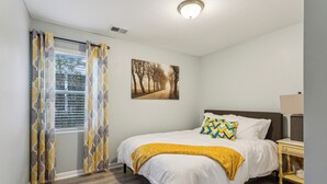 Una camera da letto con un letto, un comodino, una lampada, una finestra con tende e un quadro appeso al muro.