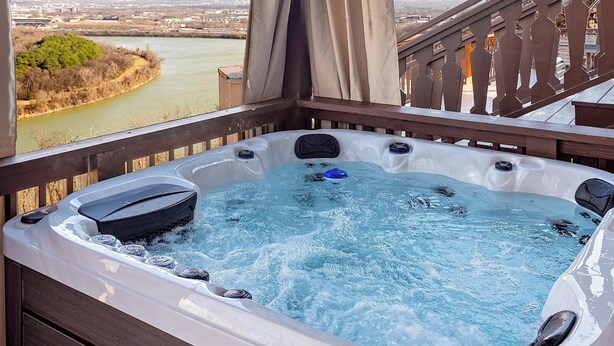 A hot tub under a wooden pergola with a view of a river and distant buildings.