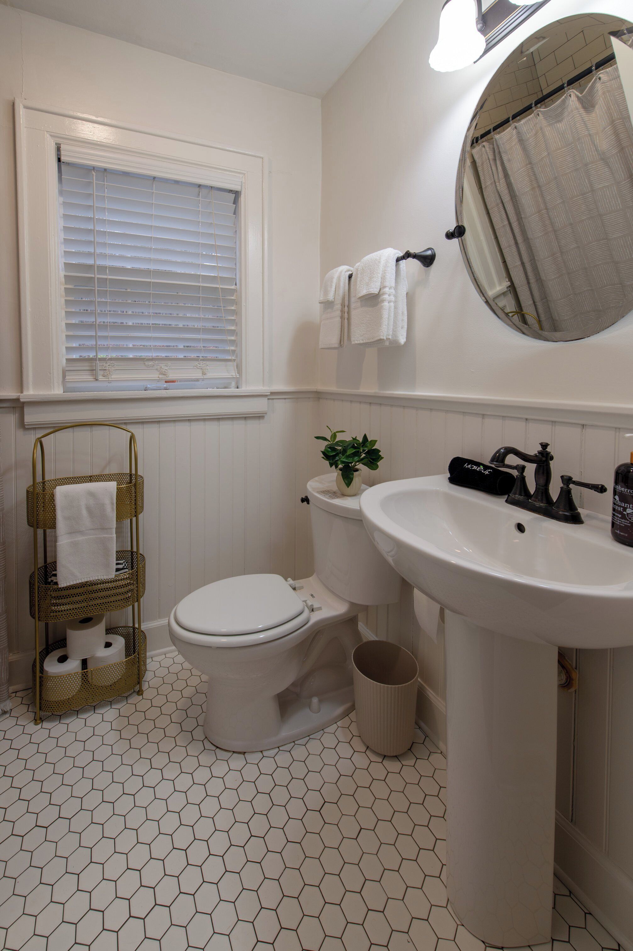 A bathroom with a toilet, sink, and window. There is a towel rack with towels, a plant, and a trash can.