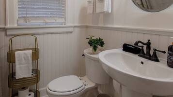 A bathroom with a toilet, sink, and window. There is a towel rack with towels, a plant, and a trash can.