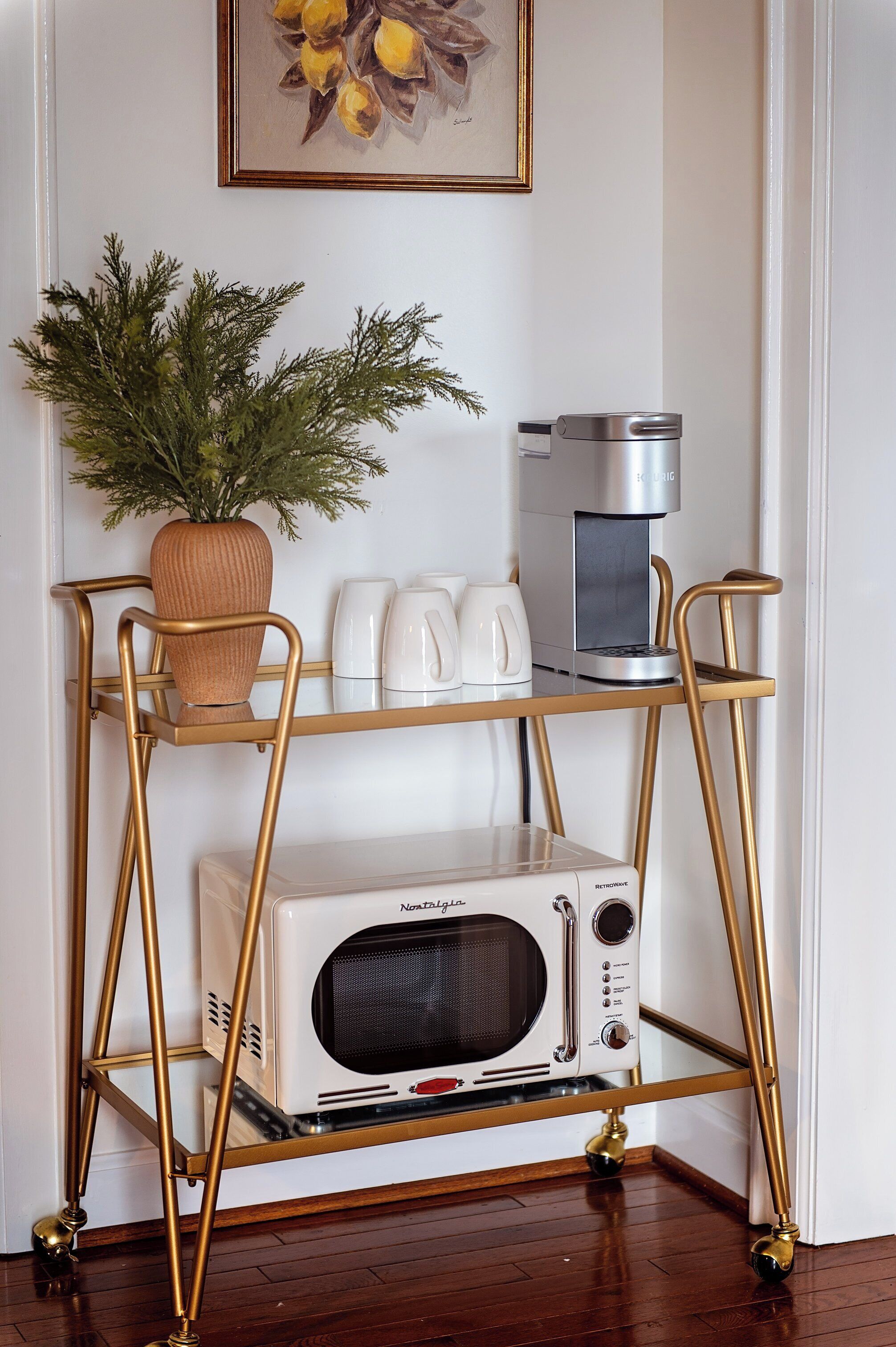 Een keukenhoek met een witte magnetron, een koffiezetapparaat en een bloempot op een gouden metalen trolley.