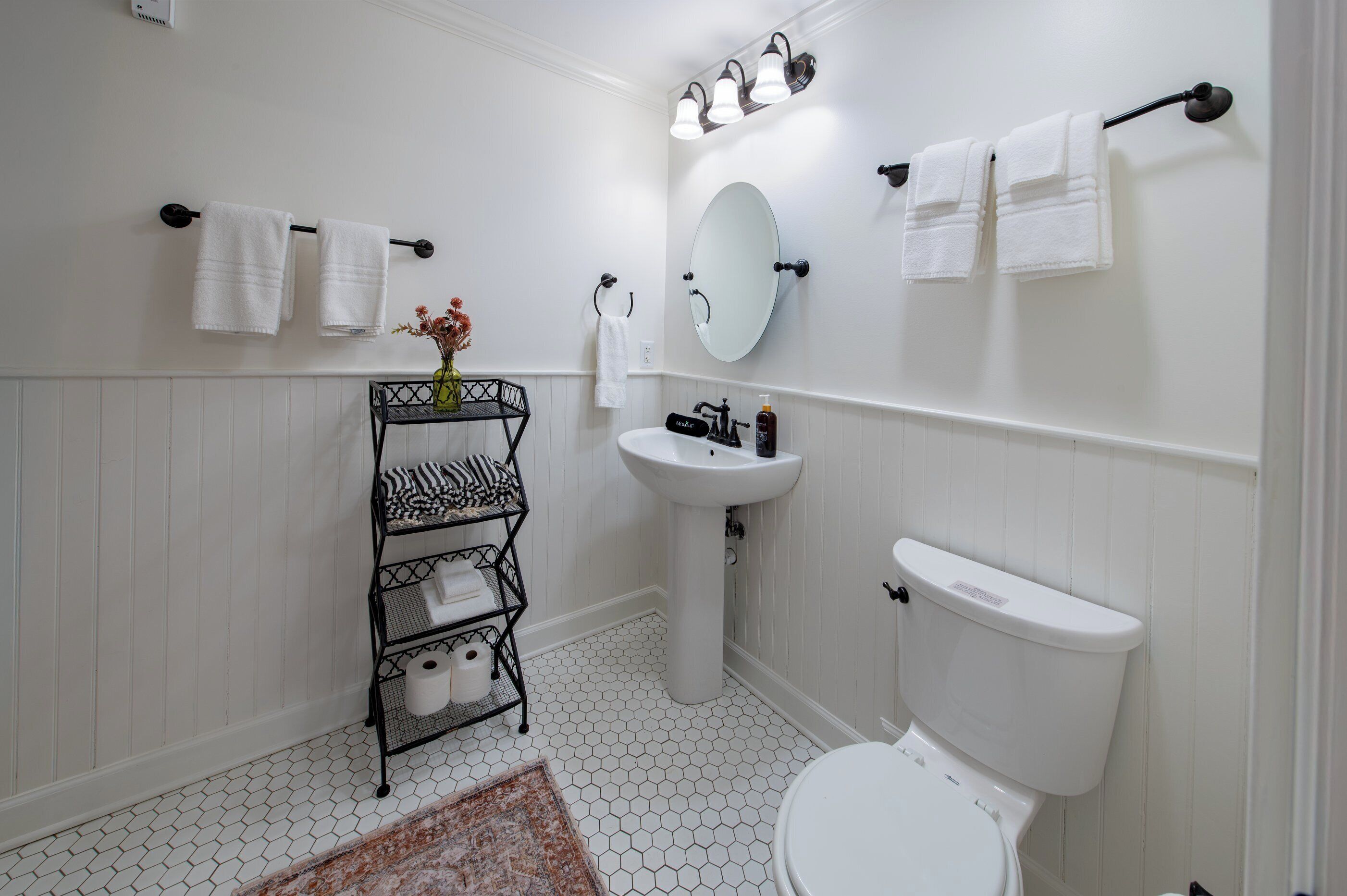 Une salle de bain avec un lavabo blanc, des toilettes et un miroir au-dessus du lavabo. Des serviettes sont suspendues sur un porte-serviettes et il y a une étagère avec des articles de toilette.