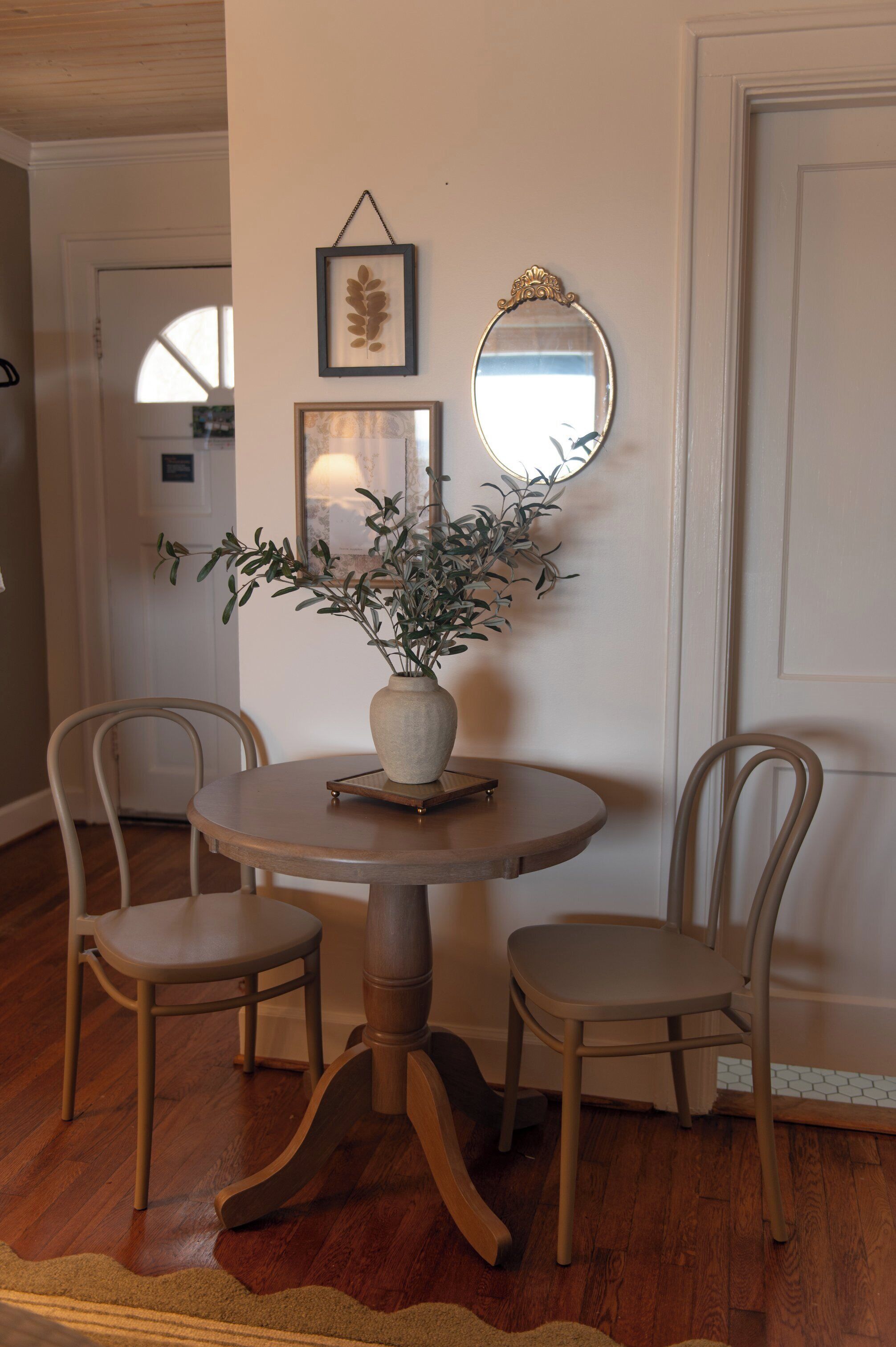 A dining area with a round table, two chairs, a vase with plants, a framed leaf artwork, and a mirror on the wall.