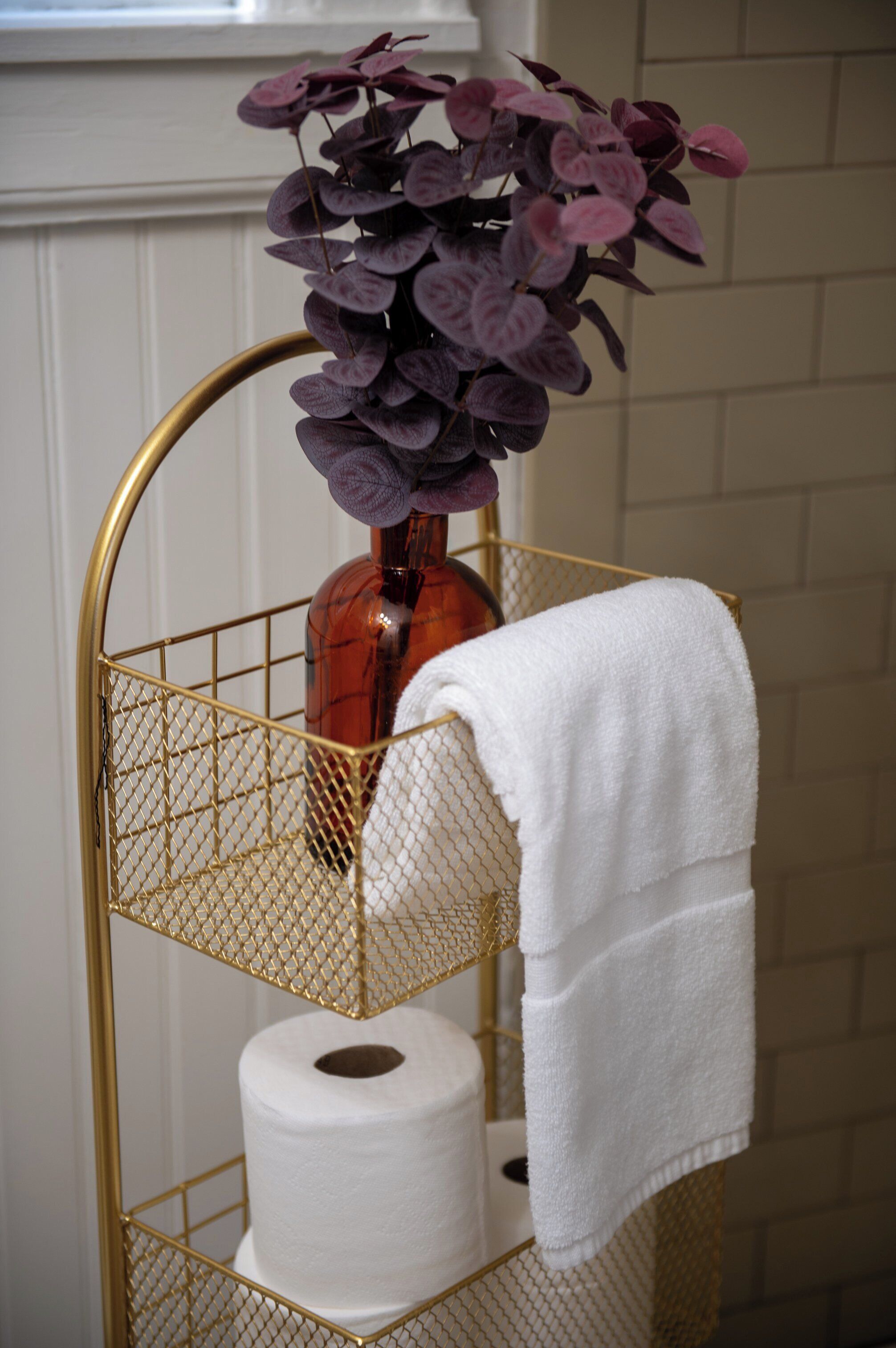 A bathroom with a gold wire basket holding a bottle, a roll of toilet paper, and a white towel.