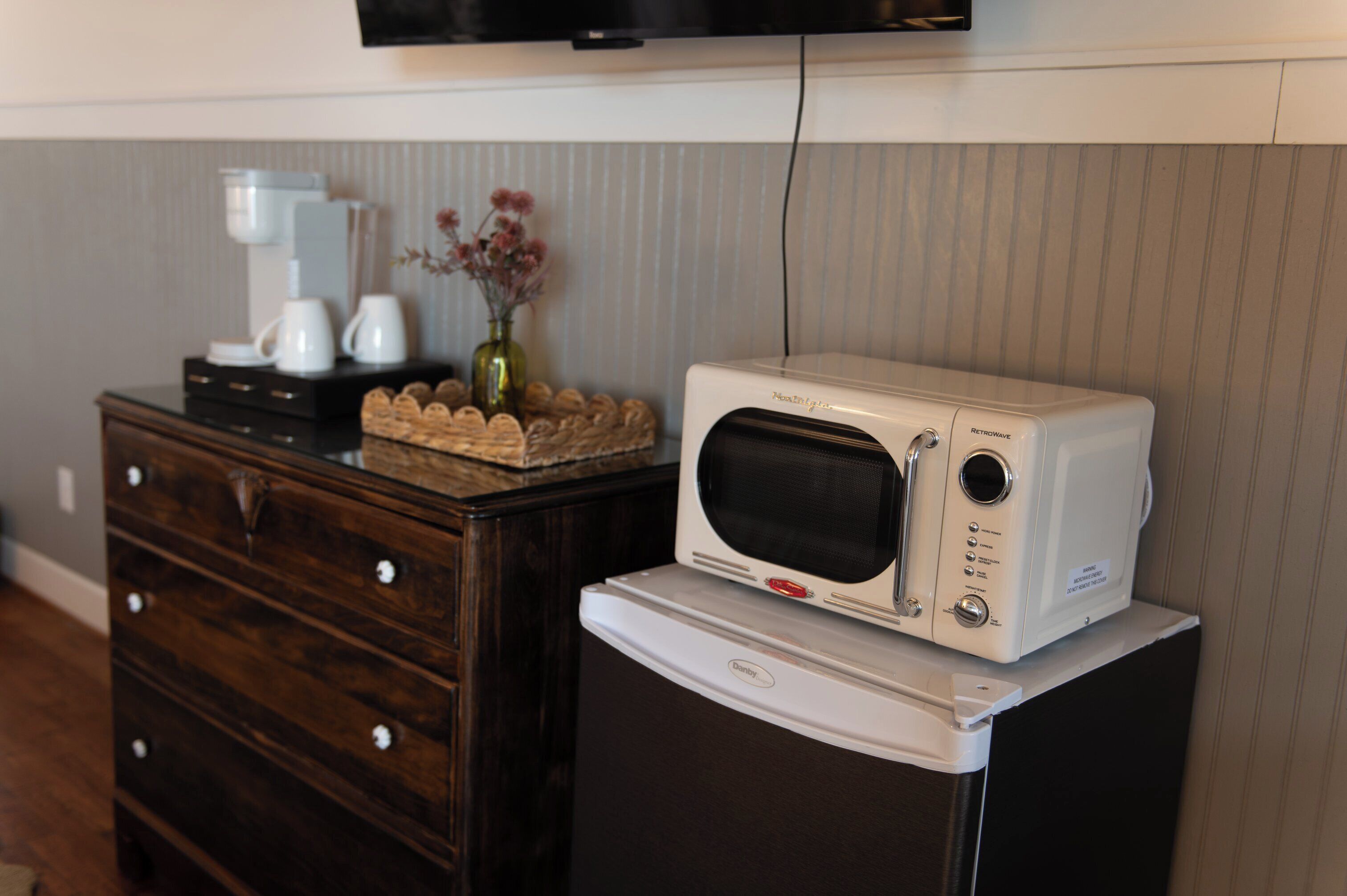 A microwave oven on top of a refrigerator, a coffee maker, and a vase with flowers on a wooden dresser.