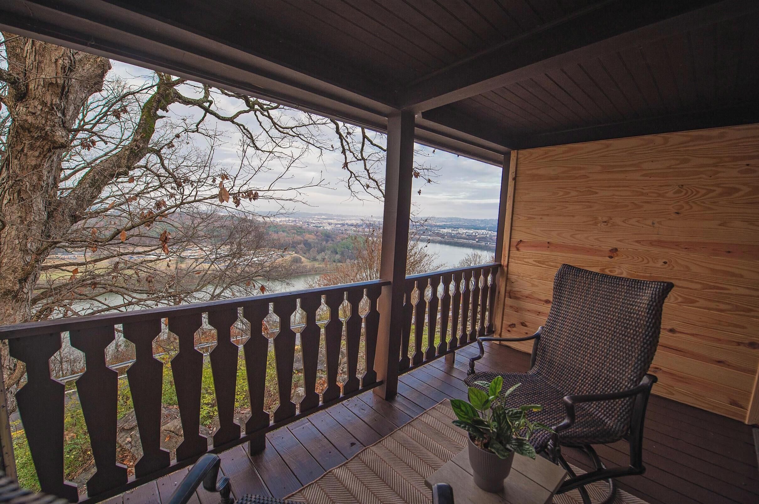 A covered porch with a view of a river and trees, a wooden wall, and a wicker chair with a potted plant.