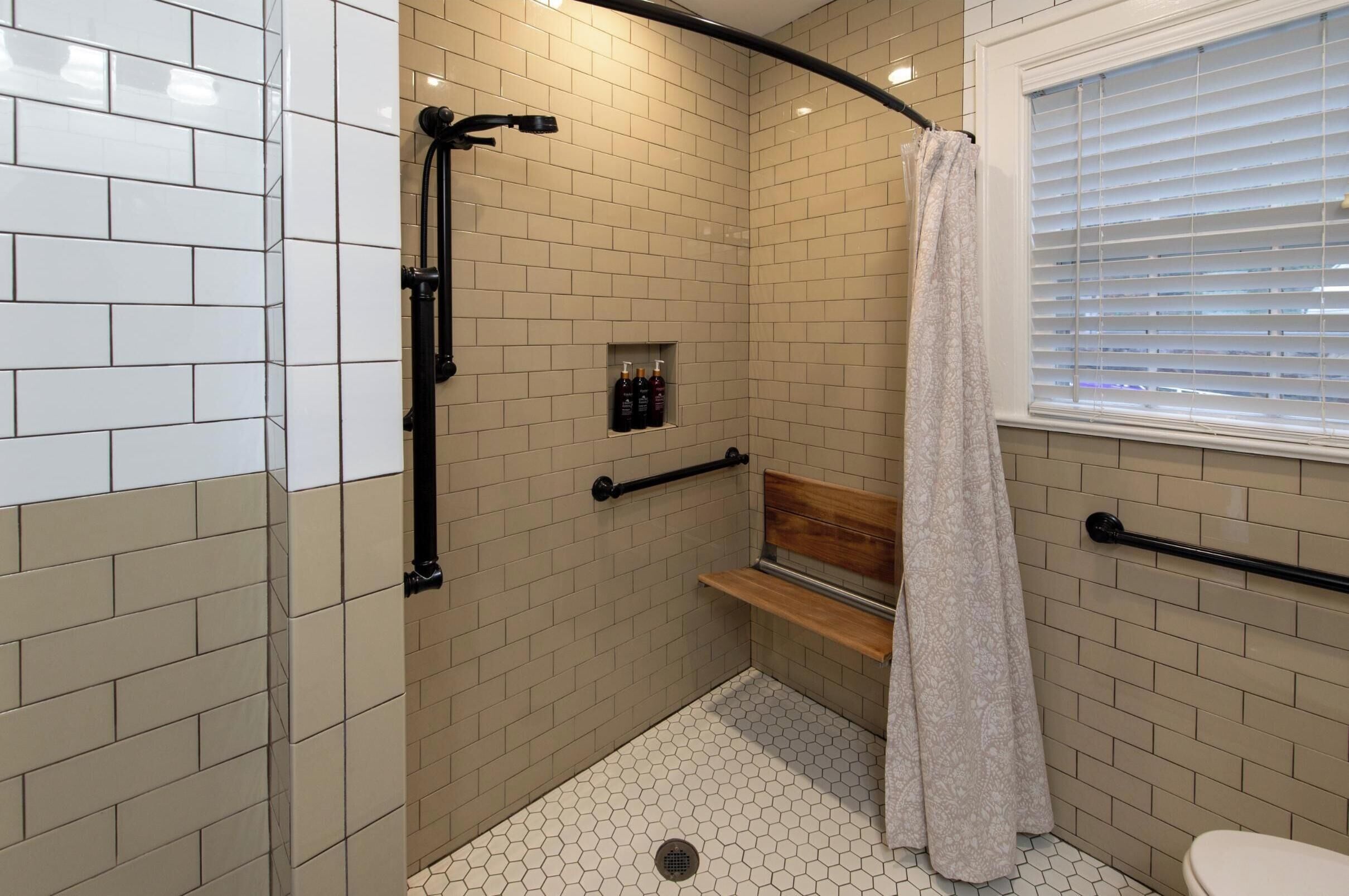 Un área de ducha pequeña y cerrada con paredes de azulejos color beige, un banco de madera y una toalla colgada en un perchero.