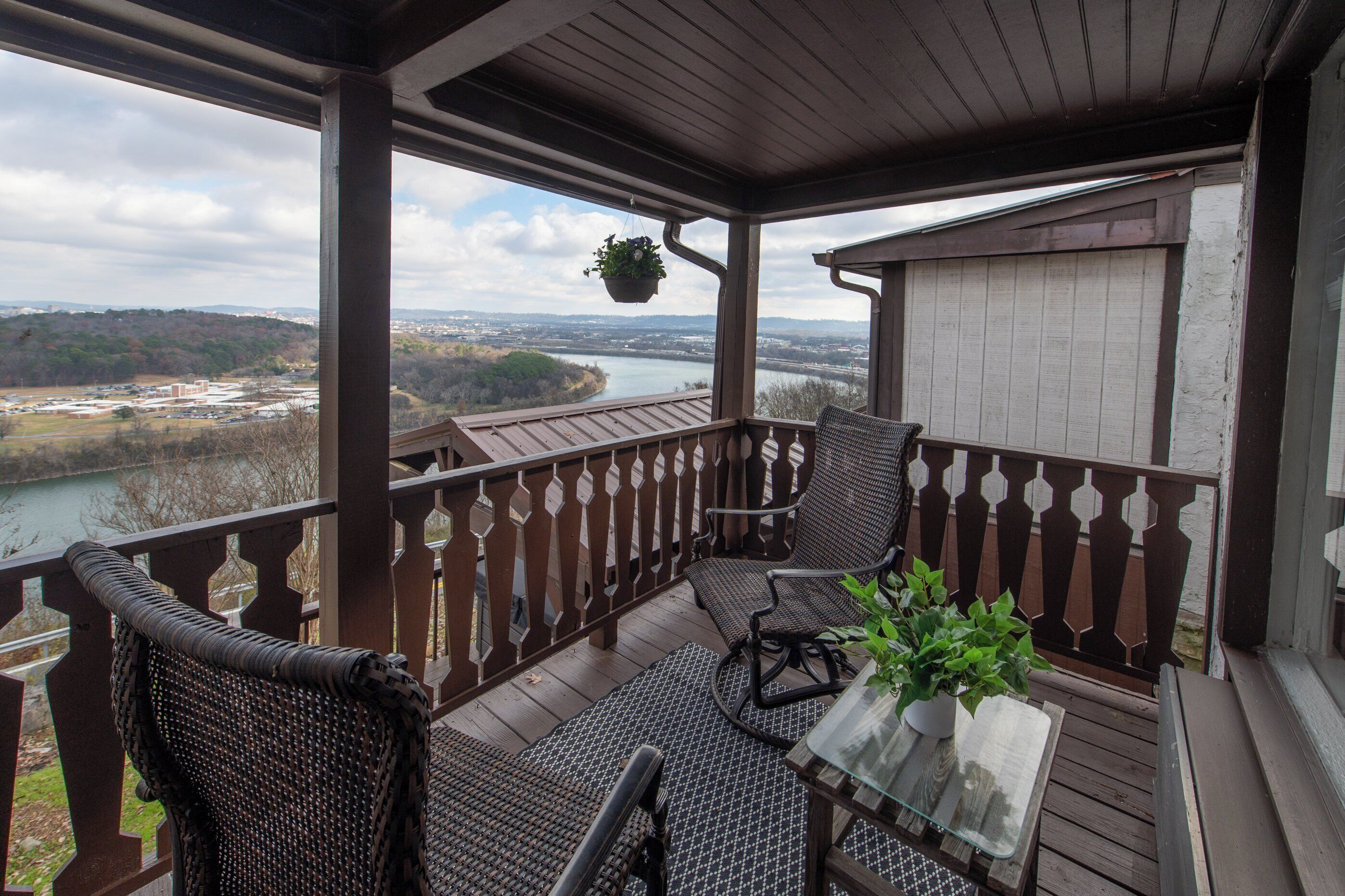 A wooden deck with wicker chairs and a table, overlooking a river and distant buildings.