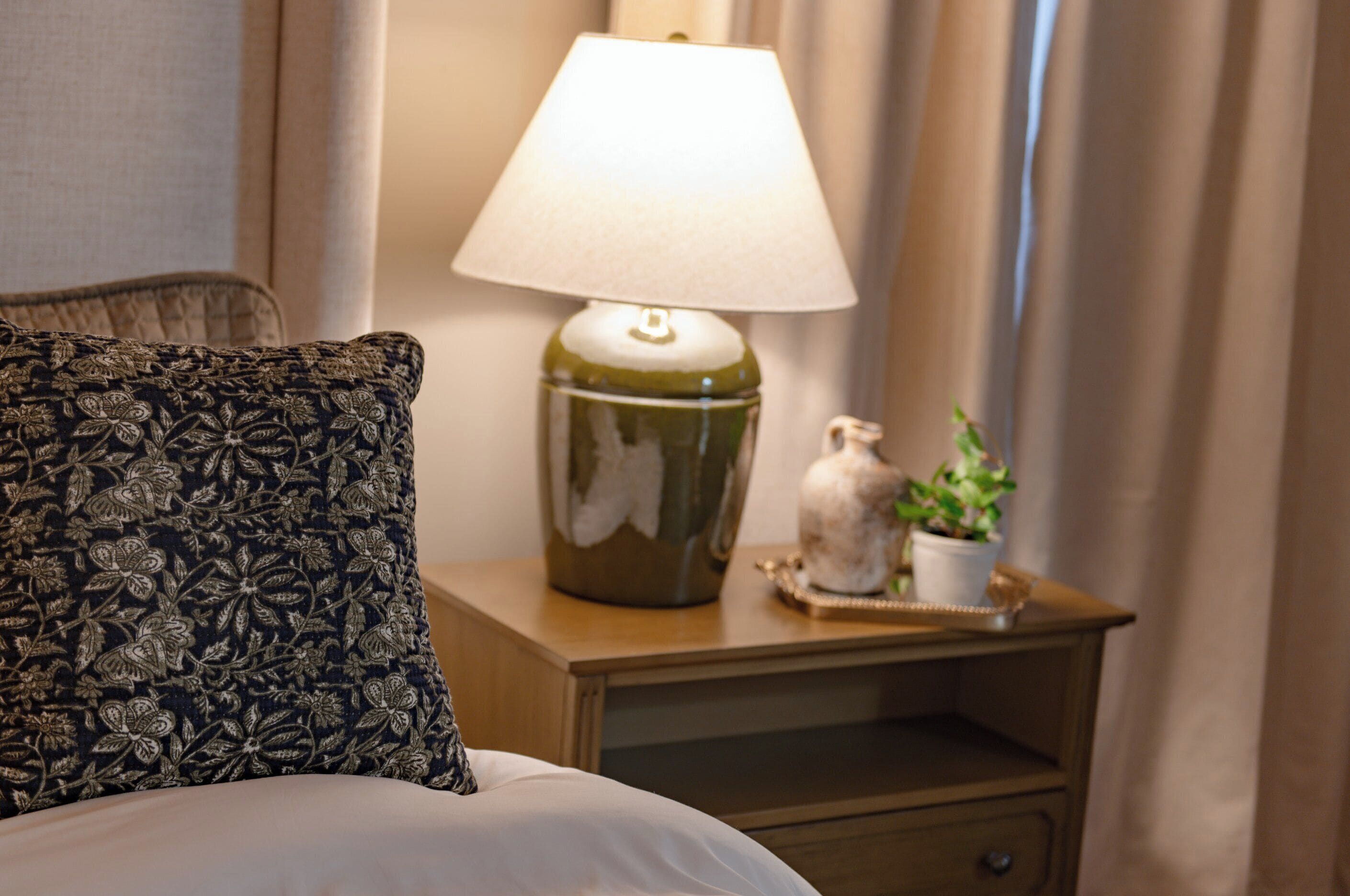 Ein ordentlich bezogenes Bett mit einem blumigen Kissen, einer Nachttischlampe, einer kleinen Vase mit Pflanze und einer Nachttischschublade.