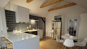 A modern kitchen with a central island, wooden beams, a dining area with white chairs, and a black tiled backsplash.