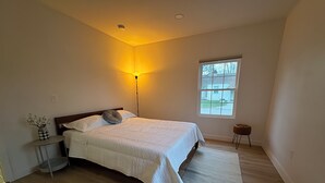 A bedroom with a bed, bedside table, lamp, window, and a small stool.