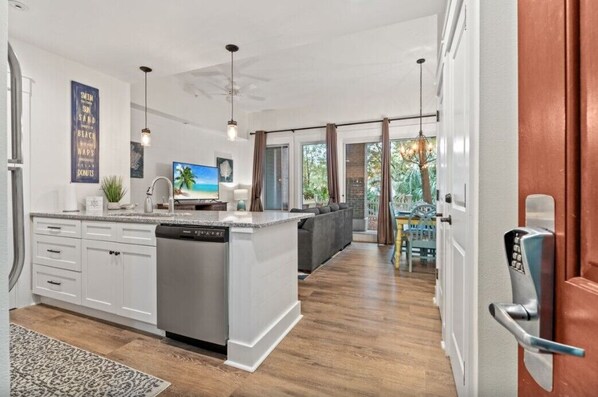 Una cocina moderna con armarios blancos, lavavajillas integrado y una isla grande. Se ve un comedor con una mesa y sillas a través de una puerta abierta.