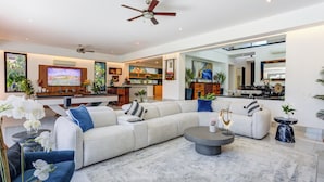 Un salón espacioso con un amplio sofá modular, una mesa de centro y una televisión. Se aprecia una cocina al fondo con una mesa de comedor y sillas.