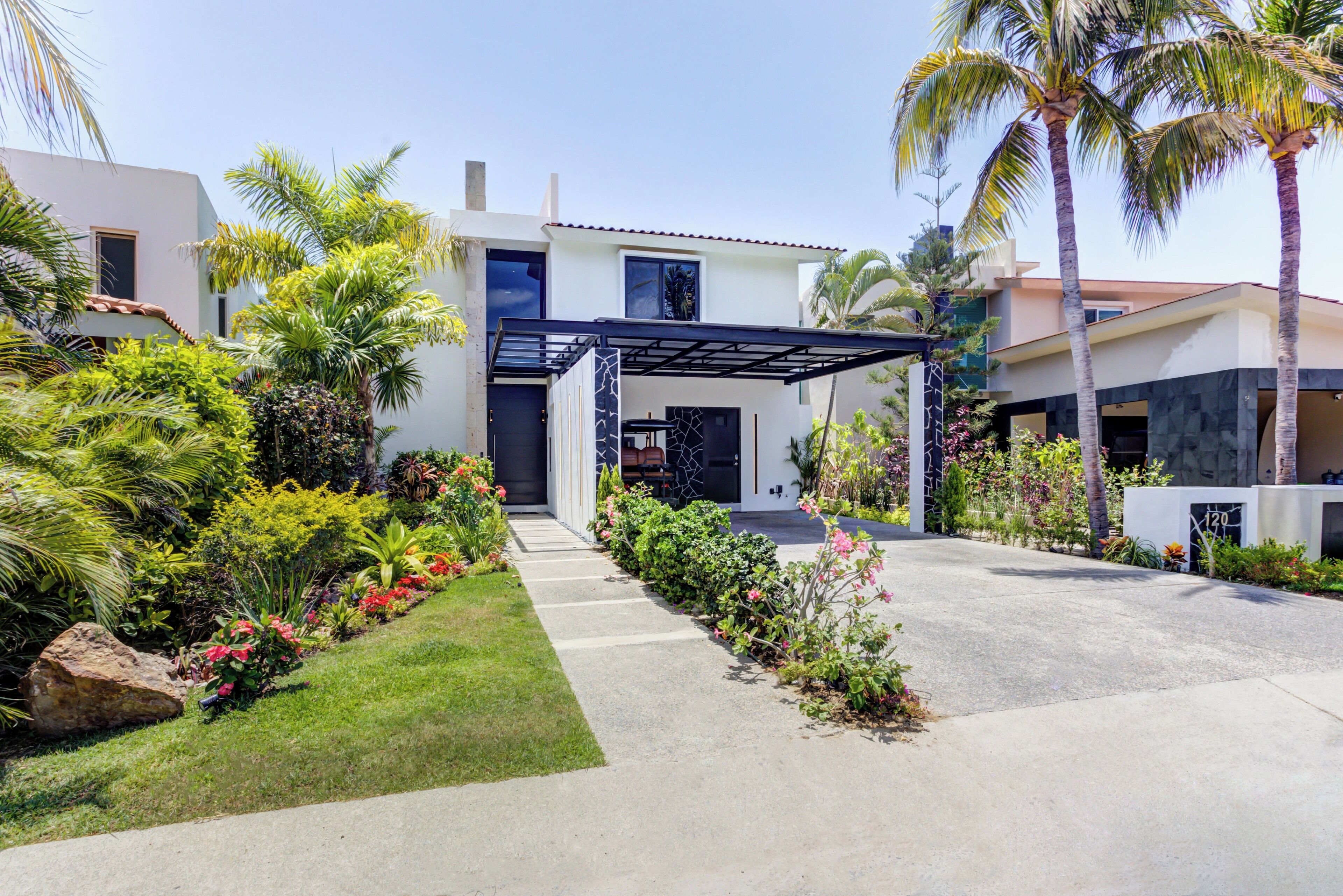 A modern house with a covered driveway, surrounded by lush greenery and palm trees.
