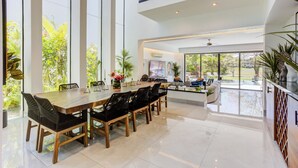 Un comedor amplio con una larga mesa de madera y sillas negras, ventanas grandes que ofrecen vistas a zonas verdes, y una cocina moderna visible a través de un vano abierto.