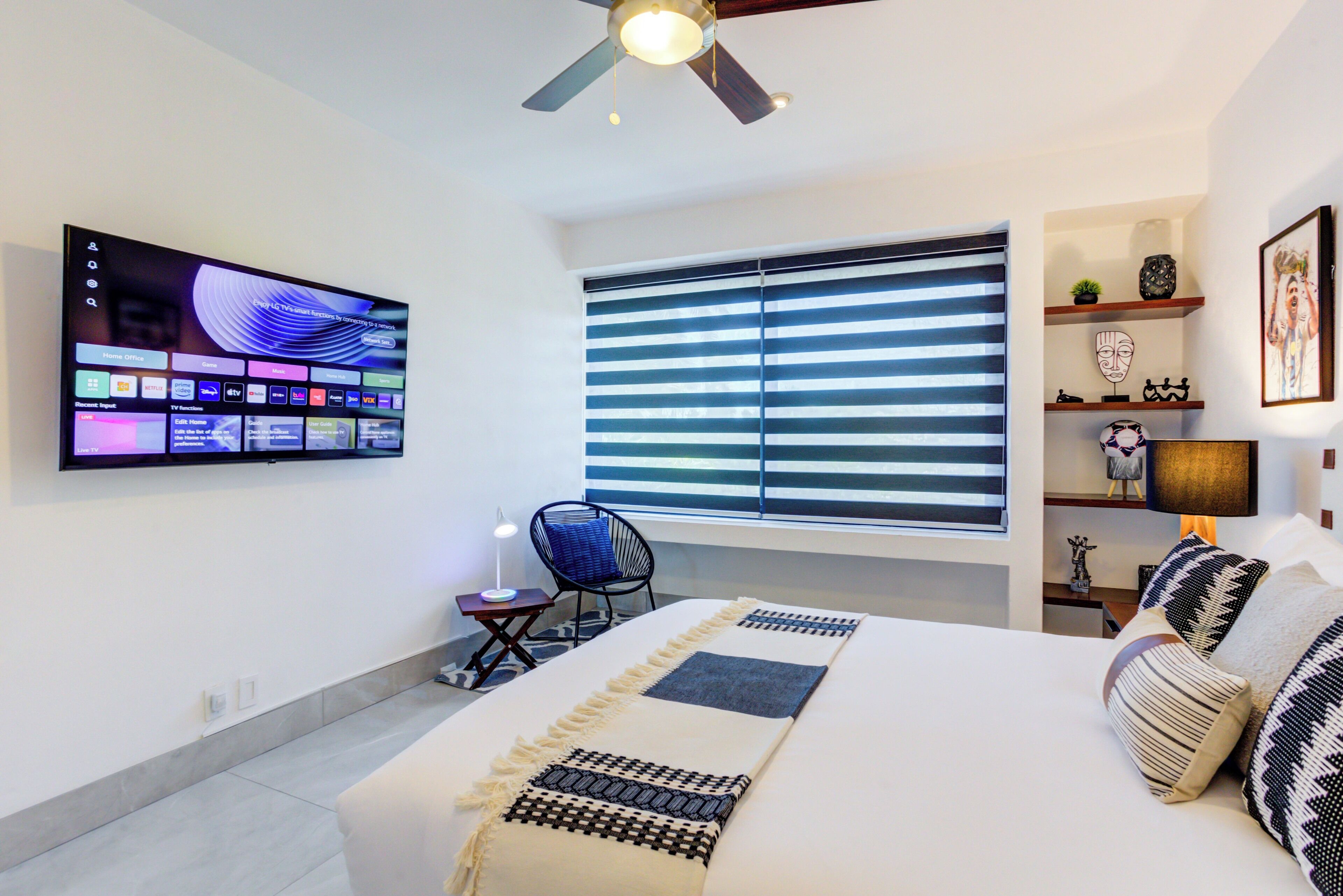 Une chambre moderne avec un grand lit, un téléviseur fixé au mur, un ventilateur de plafond et une fenêtre avec des stores.