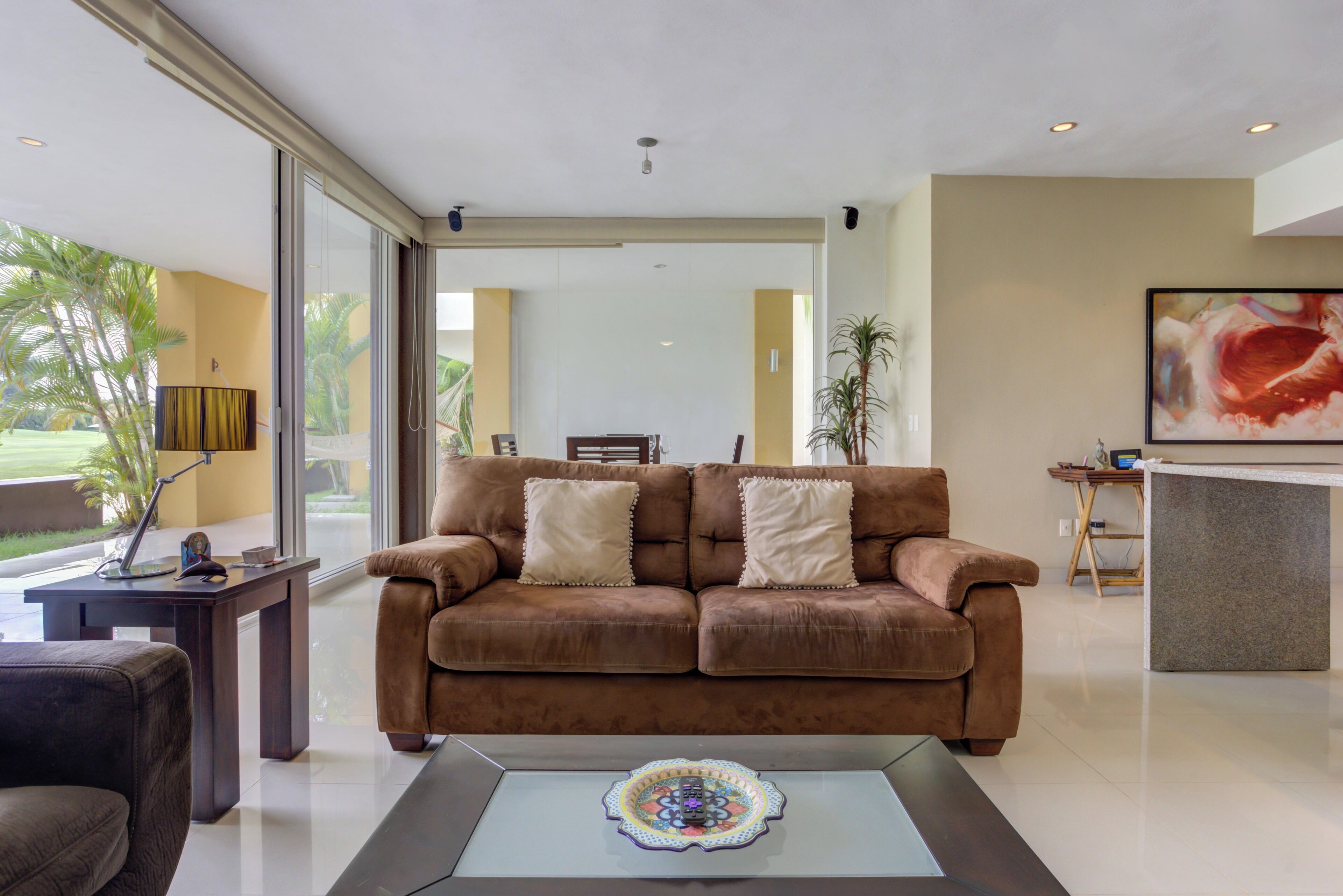 A modern living room with a brown sofa, a coffee table with a decorative item, and a large window with a view of palm trees.