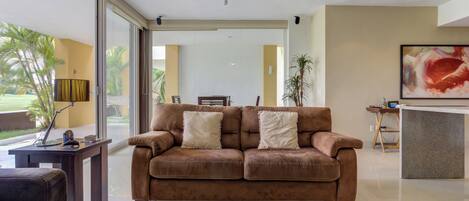 A modern living room with a brown sofa, a coffee table with a decorative item, and a large window with a view of palm trees.