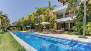 Una piscina con sillones de descanso y sombrillas, rodeada de palmeras y un edificio moderno con balcones.