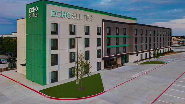 A modern hotel building with the name 'Echo Suites' prominently displayed. It features a clean, contemporary design with a mix of green and neutral colors. The building has multiple floors with a symmetrical facade, large windows, and a parking lot in front. - ECHO Suites Extended Stay by Wyndham Dallas Plano/Richardson (Plano)