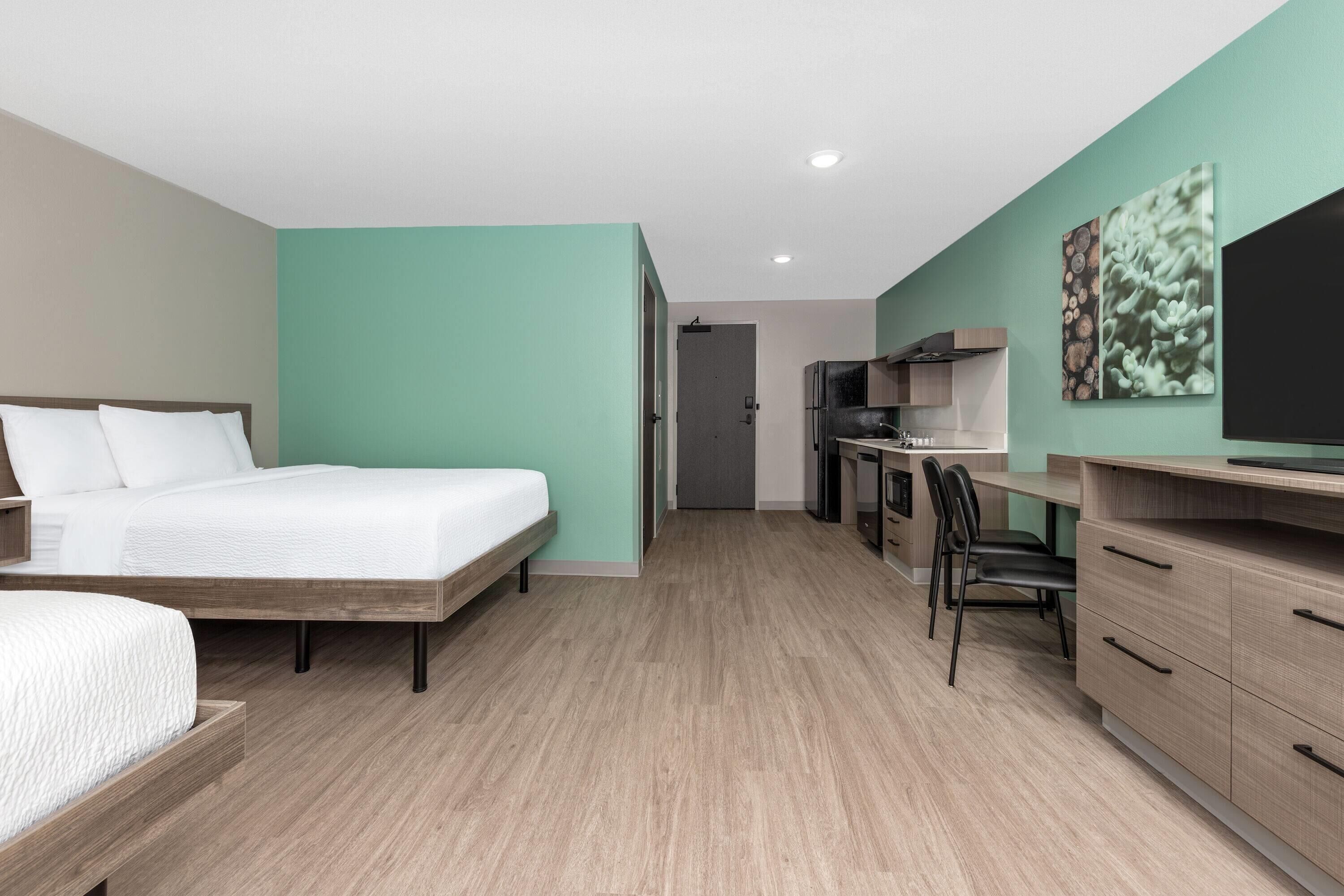 Moderni hotellihuone, jossa on sänky, työpöytä ja pieni keittiö.