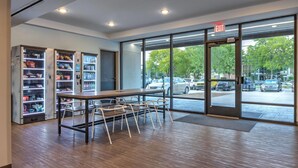 A modern indoor space with a wooden table and chairs, vending machines, and large windows offering a view of parked cars and trees outside. - ECHO Suites Extended Stay by Wyndham Dallas Plano/Richardson (Plano)