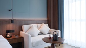 Habitación de hotel con sofá, mesita de noche con teléfono, mesa con tetera y taza, y ventana con cortinas.