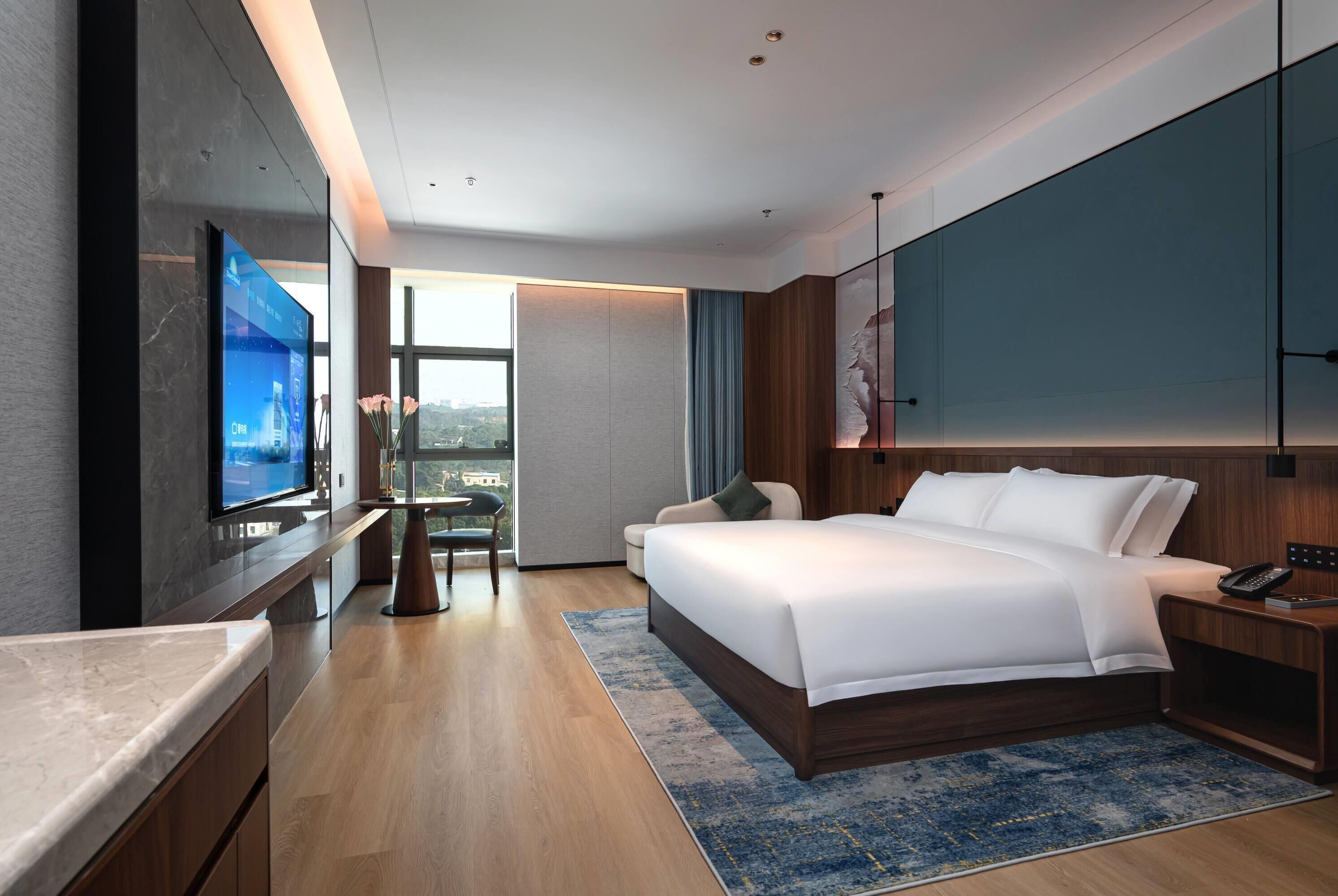A modern hotel room with a large bed, a TV mounted on the wall, a desk, and a view of the city.