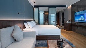 Habitación de hotel moderna con una cama grande, un sofá, un televisor y un baño visible a través de una puerta de cristal.
