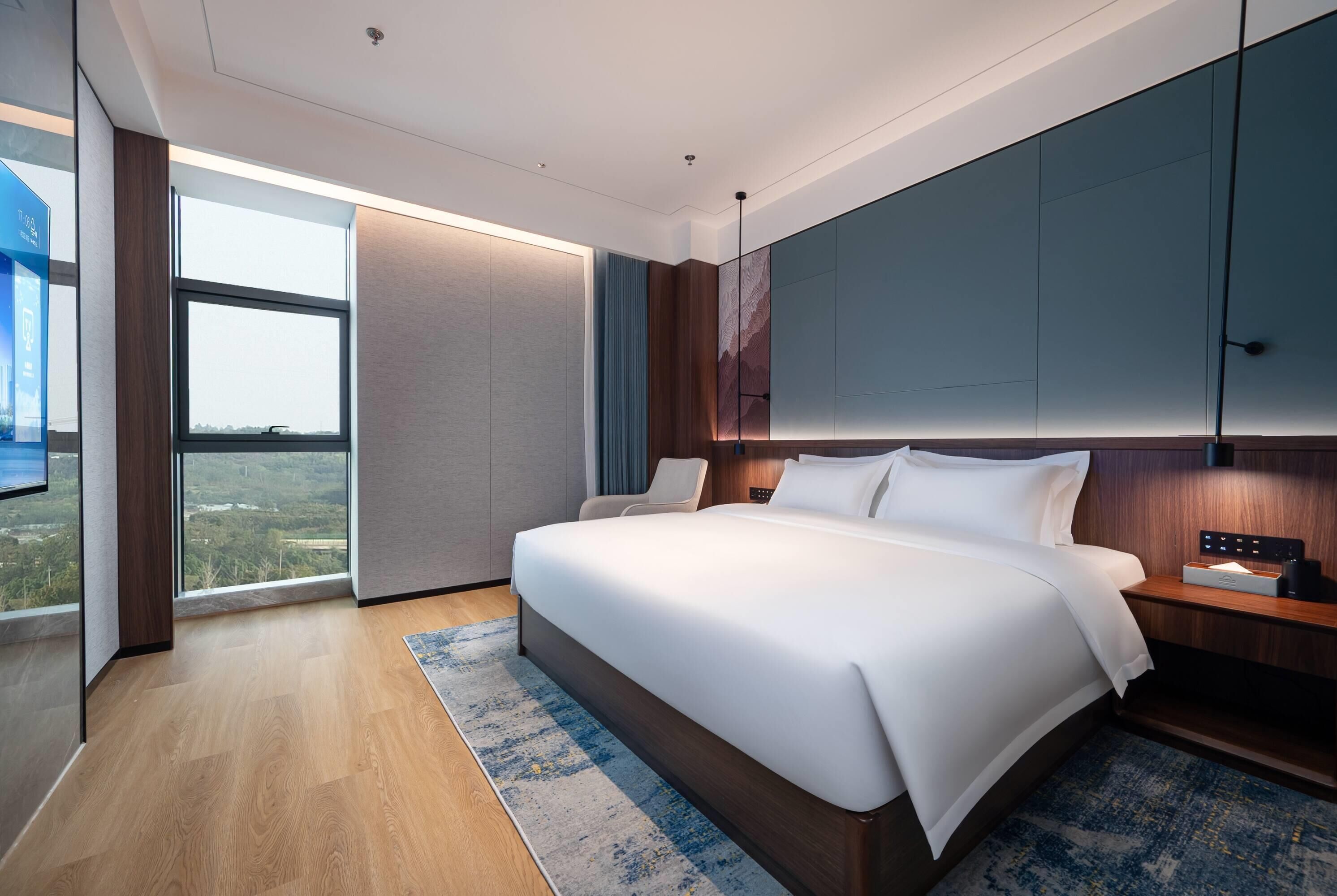 Quarto de hotel moderno com uma cama grande, uma mesinha de cabeceira, uma TV e vista para a cidade.