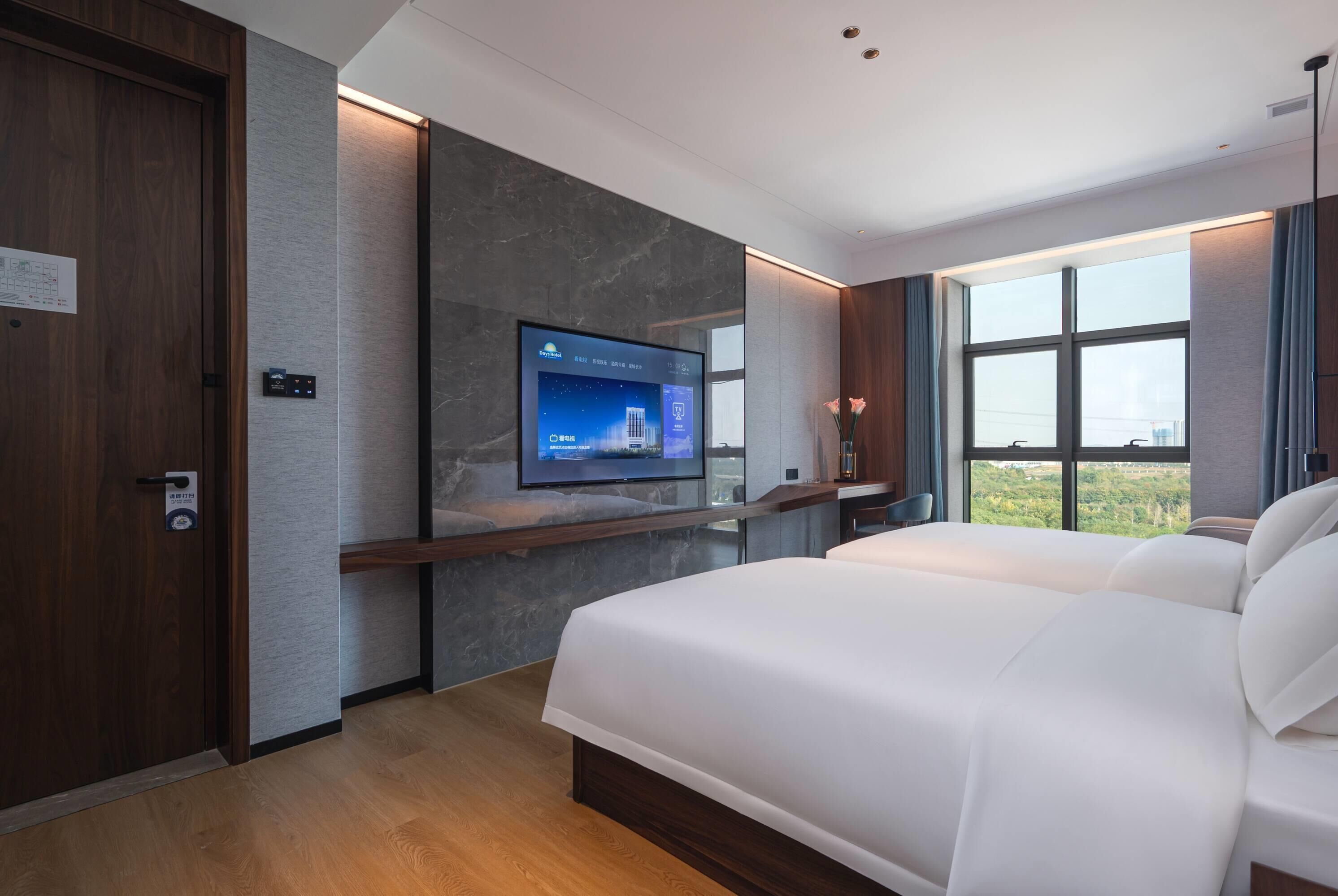 A modern hotel room with a large bed, a mounted TV, a wooden door, and a window with a city view.