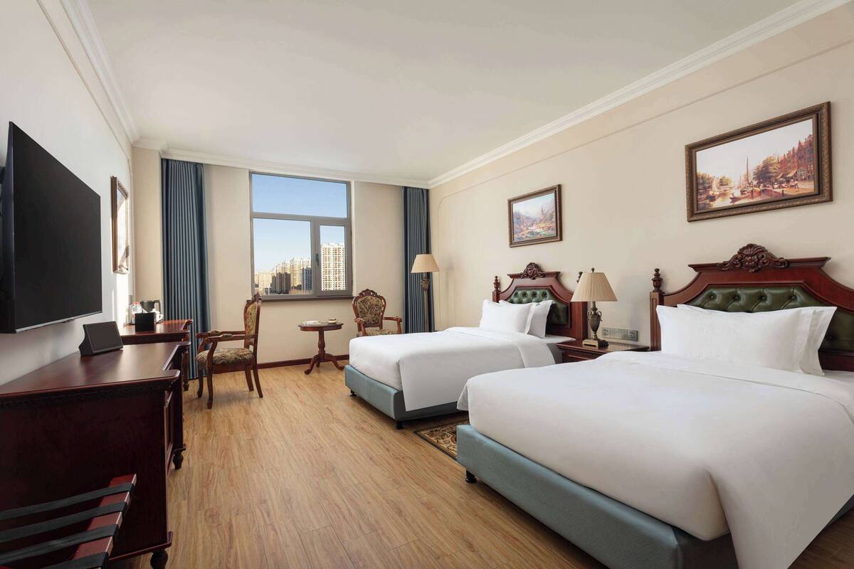 Habitación de hotel con dos camas, un escritorio, un televisor, una ventana con vistas a la ciudad y un cuadro en la pared.