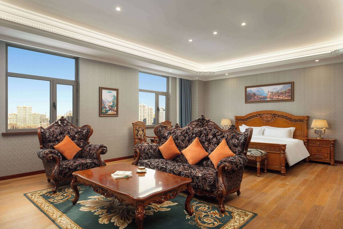 Habitación de hotel con una cama grande, dos sillones decorados, una mesa de centro de madera y vistas a la ciudad a través de amplios ventanales.