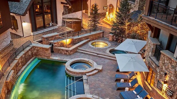 Un área de piscina en la azotea con varios jacuzzis y sillones de descanso, rodeada de muros de piedra y una escalera de piedra que conduce a un nivel superior.