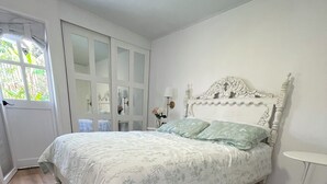 A bedroom with a wooden floor, a white bed with a floral bedspread, a white headboard, and a small bedside table with a lamp.