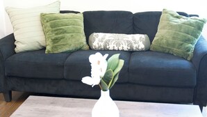 Um sofá azul escuro com almofadas verdes e estampadas, um vaso branco com uma flor sobre uma mesa de centro de madeira e duas obras de arte abstratas na parede.