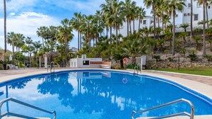 A clear blue swimming pool with metal railings, surrounded by palm trees and a white building in the background. - Las Palmas 20c - El Oasis de Capistrano (Nerja)