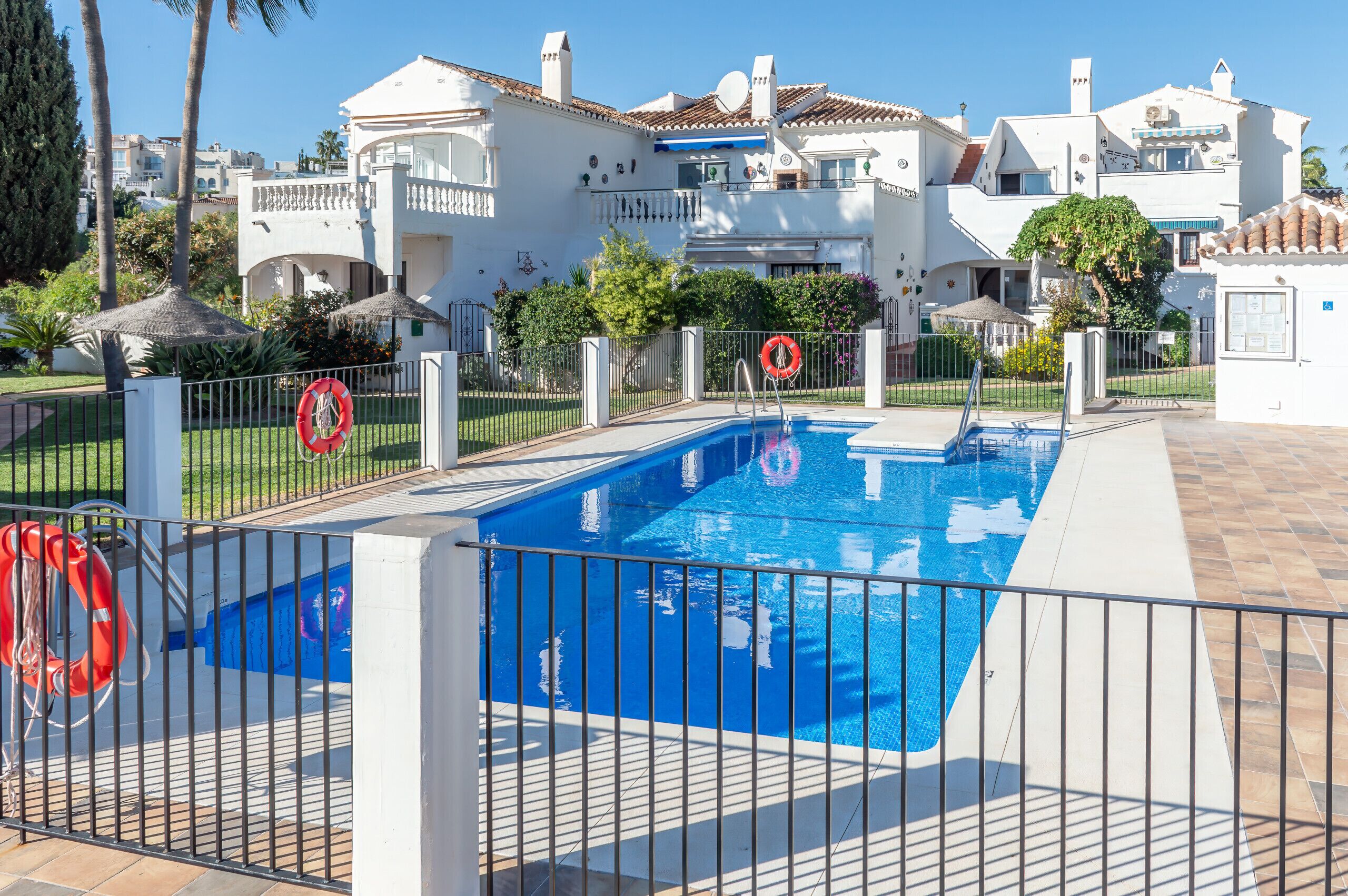 Una piscina con un flotador salvavidas, rodeada por una valla, y un edificio blanco con techo de tejas al fondo.
