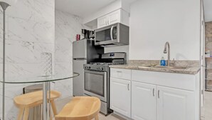 A compact kitchen with white cabinets, a stainless steel oven, a microwave, and a sink. There is a glass table with two stools. - Bahama Mama Studio Suite (Atlanta)