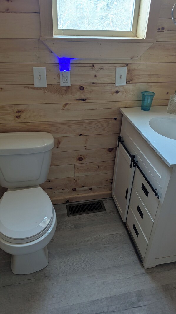 Een kleine, functionele badkamer met een houten wand, een wit toilet, een wastafel met spiegel en een raam boven de wastafel.