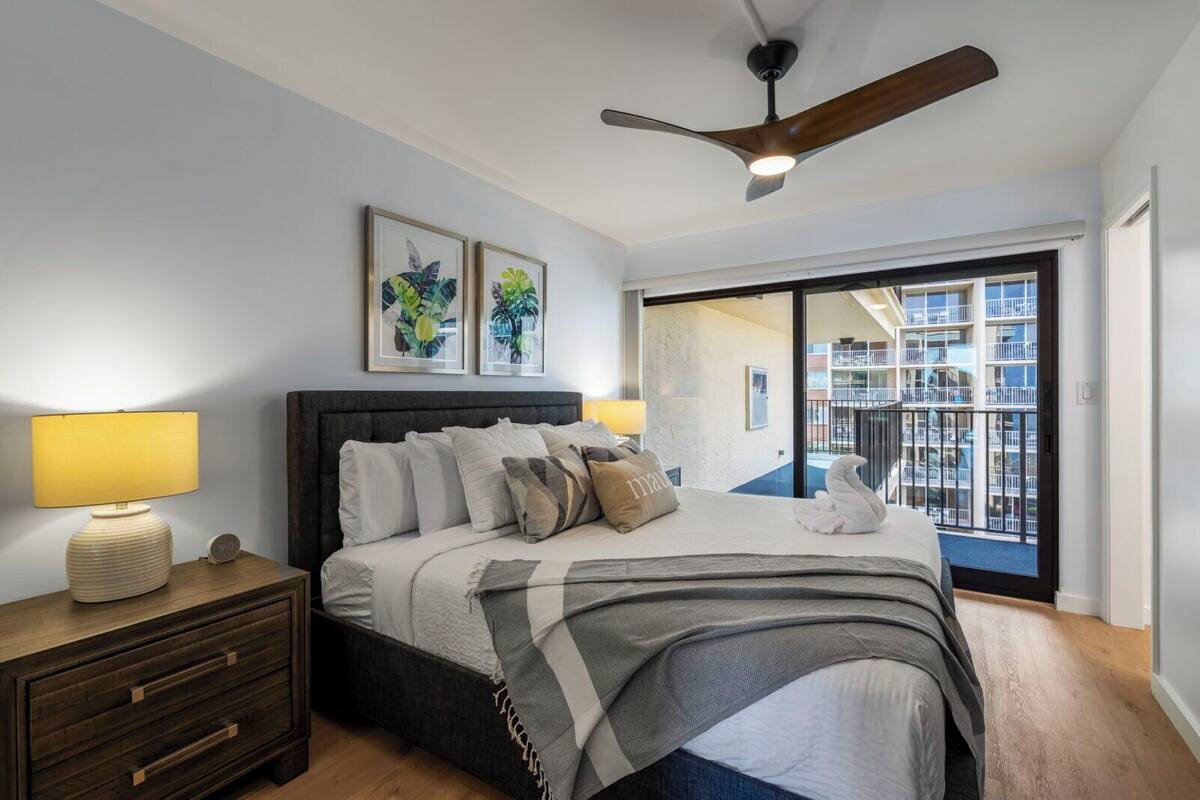 A modern bedroom with a bed, bedside lamps, a ceiling fan, and a large window with a balcony view.