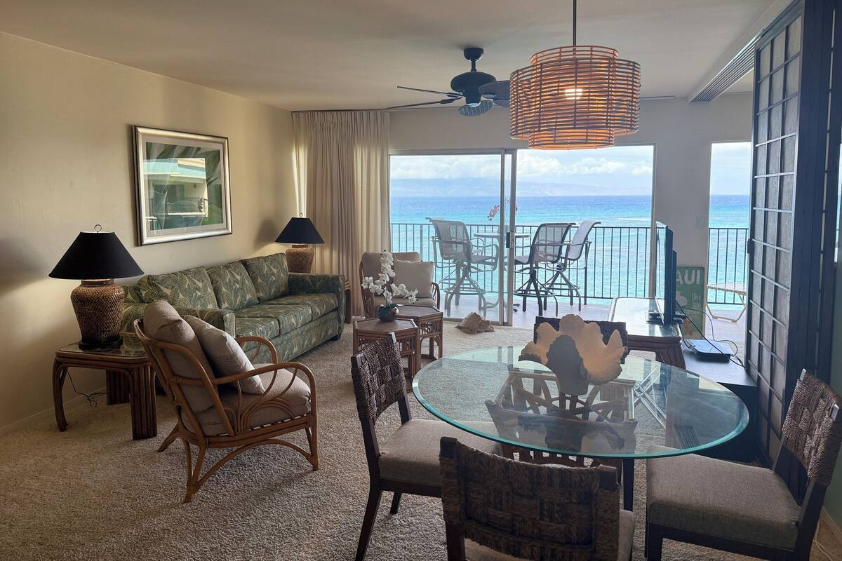 A living room with a sofa, chairs, a glass table, and a view of the ocean.