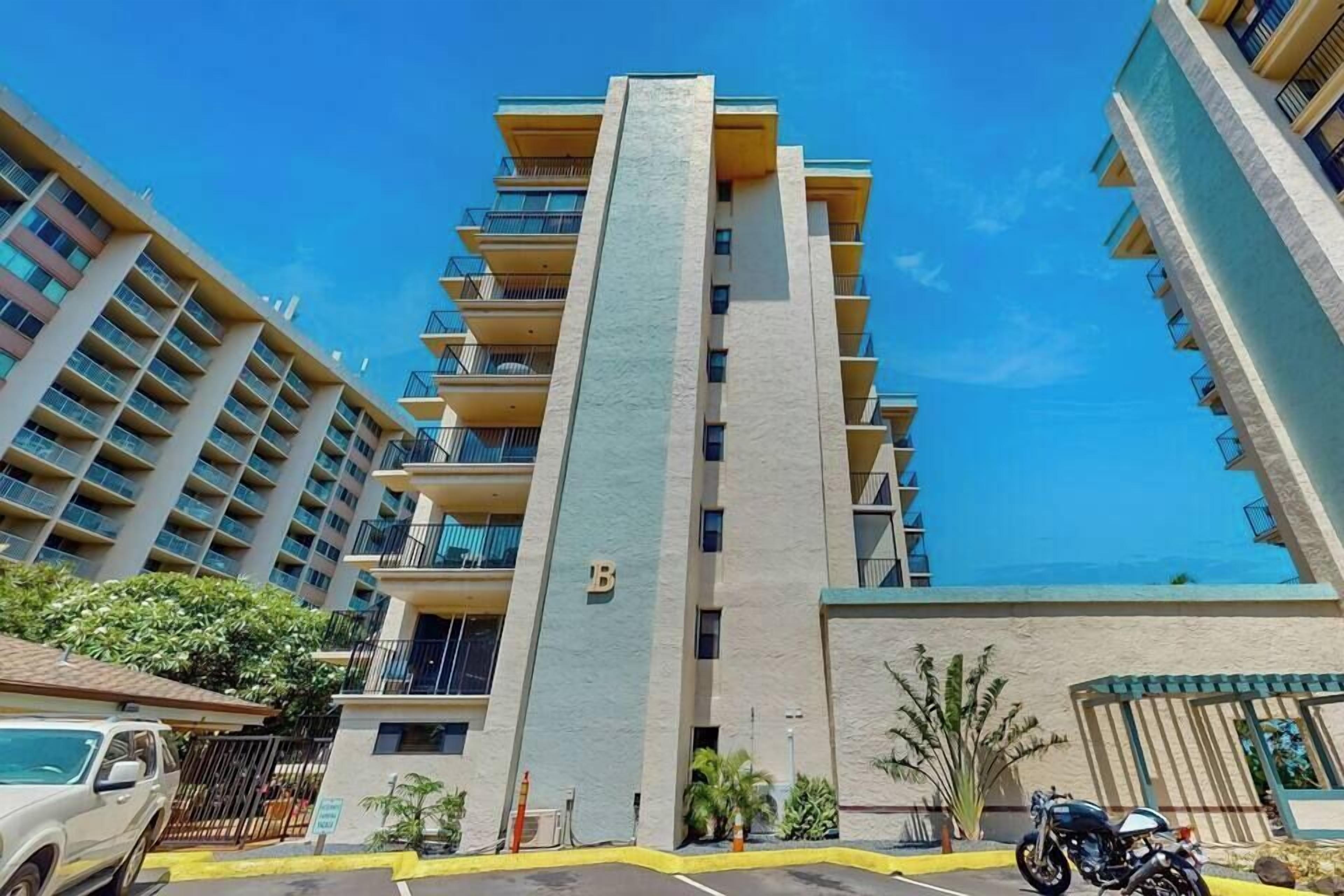 A multi-story residential building with balconies, a parking area, and a motorcycle parked in front.