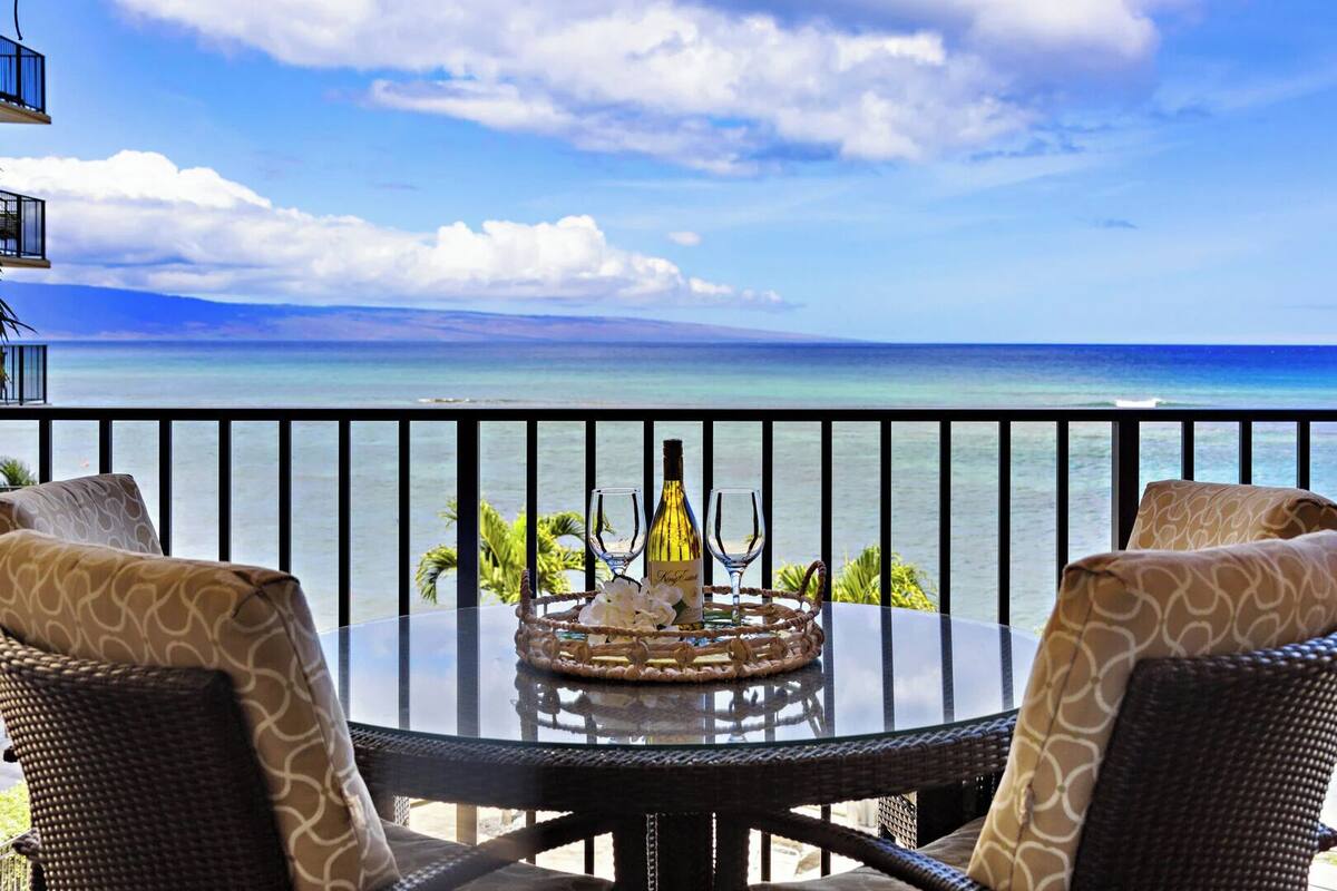 A balcony with a glass table, two wicker chairs, a bottle of wine, and a glass on the table.