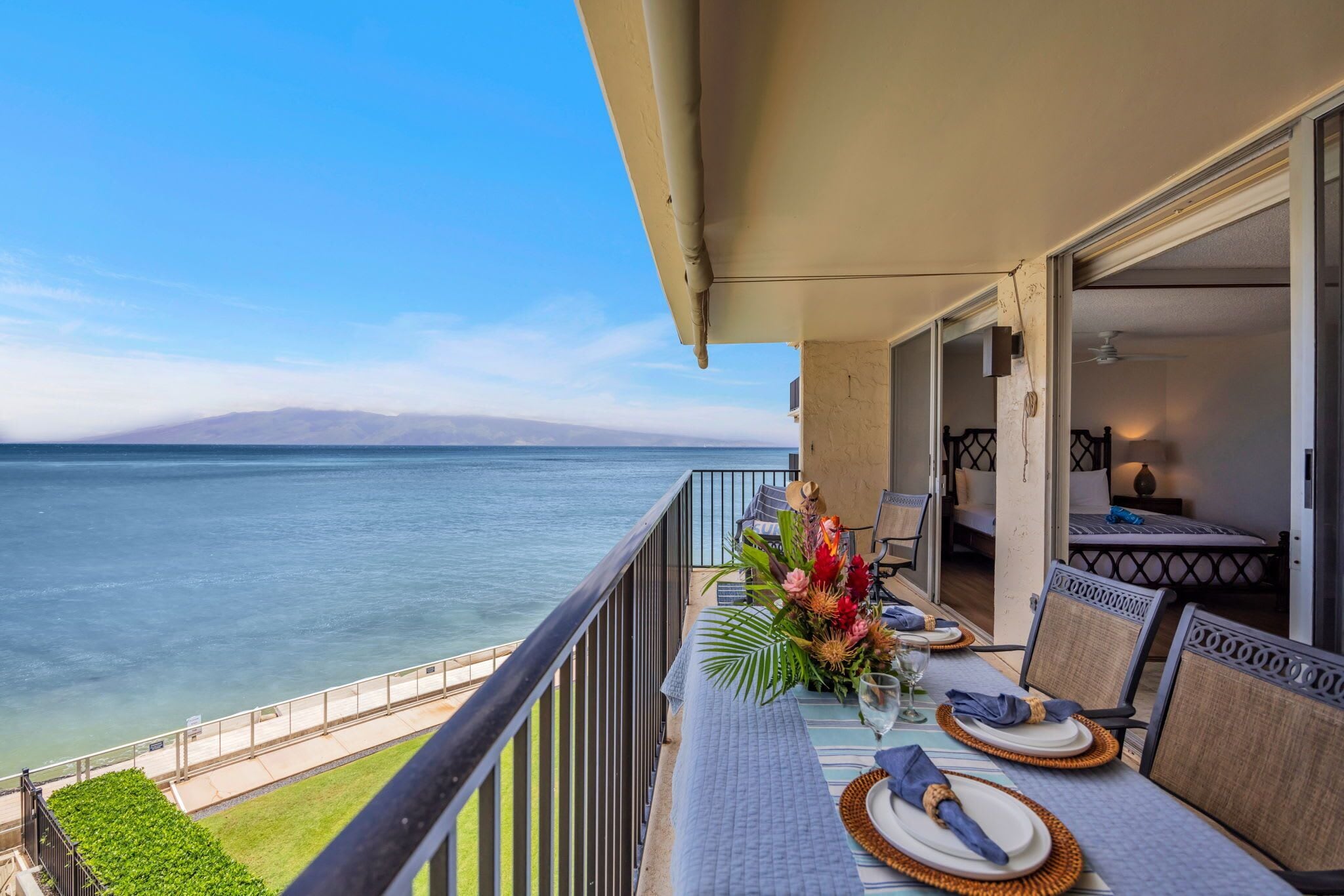 A balcony with a view of the ocean, a dining table set with plates and a vase of flowers, and a seating area with chairs.
