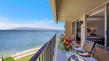 A balcony with a view of the ocean, a dining table set with plates and a vase of flowers, and a seating area with chairs.