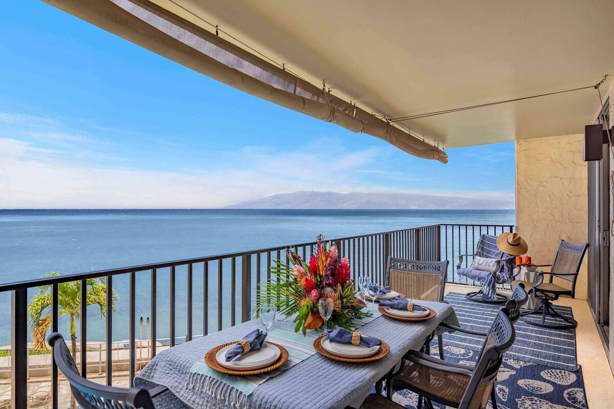 A balcony with a table set for dining, chairs, a decorative vase, and a view of the sea.