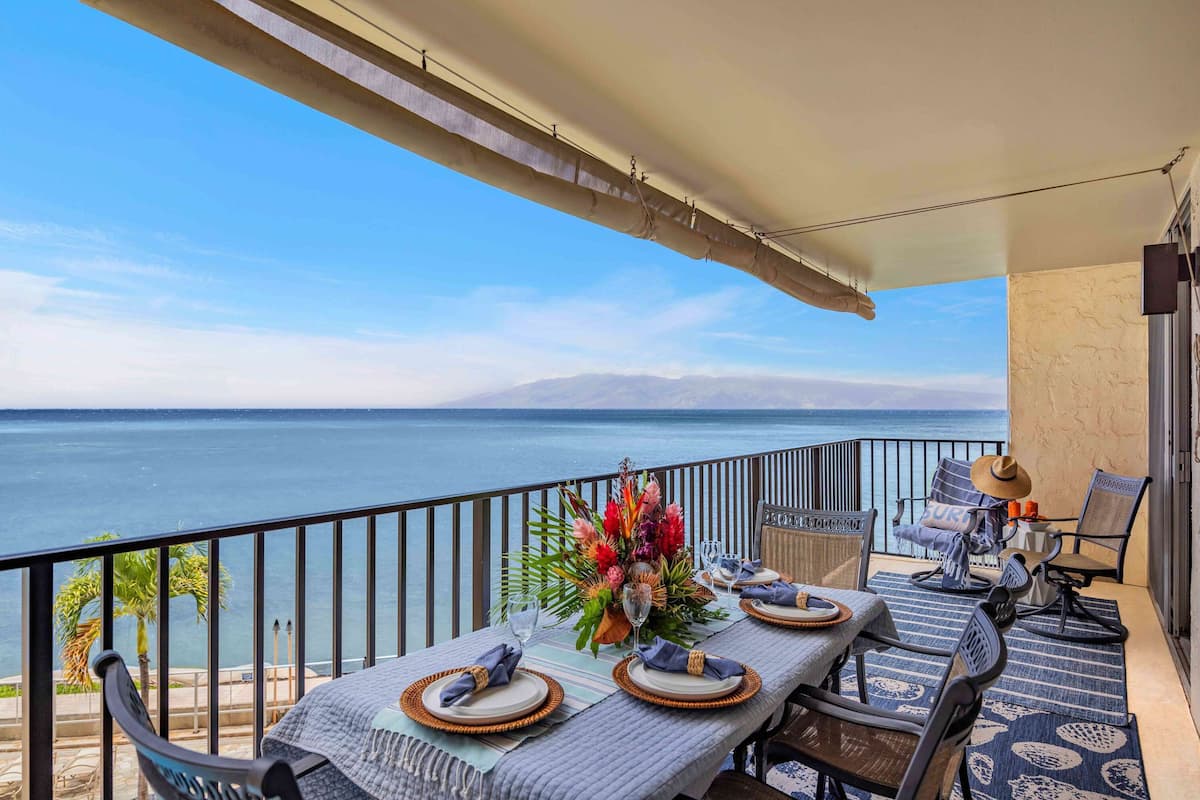 A balcony with a table set for dining, chairs, a decorative vase, and a view of the sea.