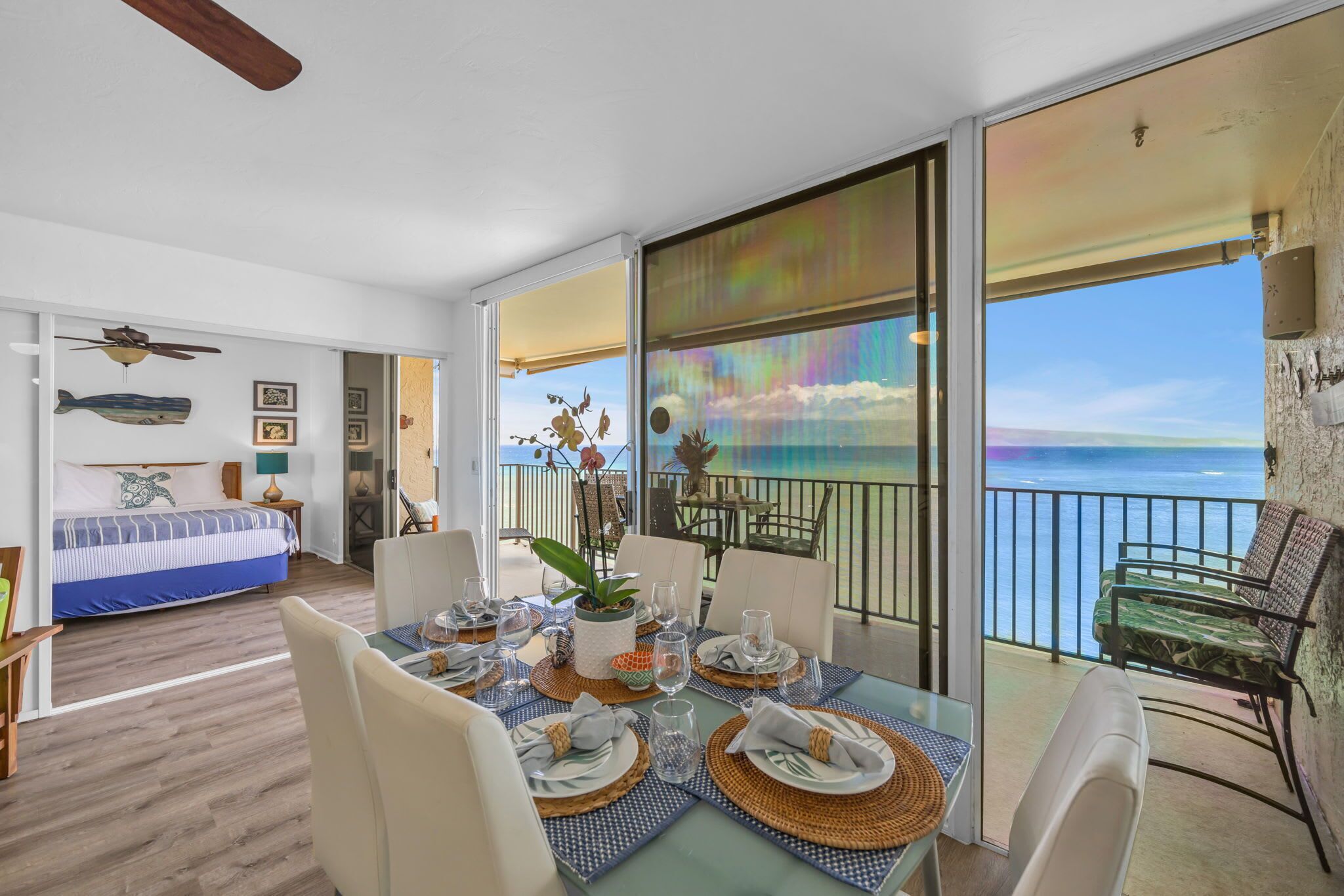 Un coin repas avec une table dressée pour quatre personnes, un lit recouvert d’une couvre-lit à rayures bleues et blanches, et un balcon offrant une vue sur l’océan.