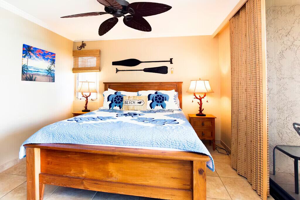 A bedroom with a wooden bed, two wall-mounted lamps, a ceiling fan, a beach-themed pillow, and a beach-themed wall hanging.